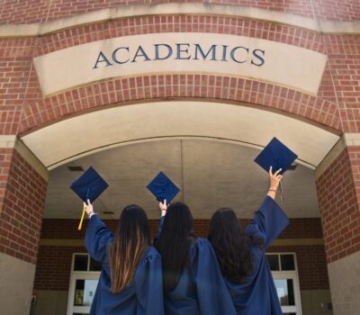 Three female students holding their mortarboards aloft underneath arch with 'ACADEMICS' inscribed into the stone.
