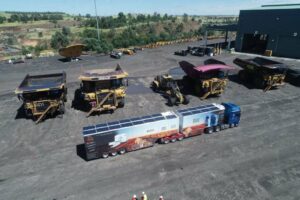The Dust Bus - a large lorry with a red desert decal and MRI painted on it's side, next to a group of mining drucks in an open cut coal mine