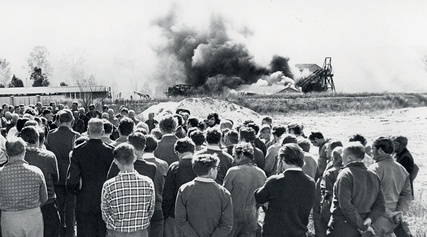 A group of people stand in front of a coal mine smouldering
